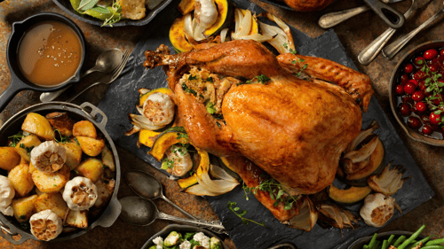  Roasted stuffed turkey on slate platter surrounded by roasted garlic bulbs, gravy, and potatoes on table 